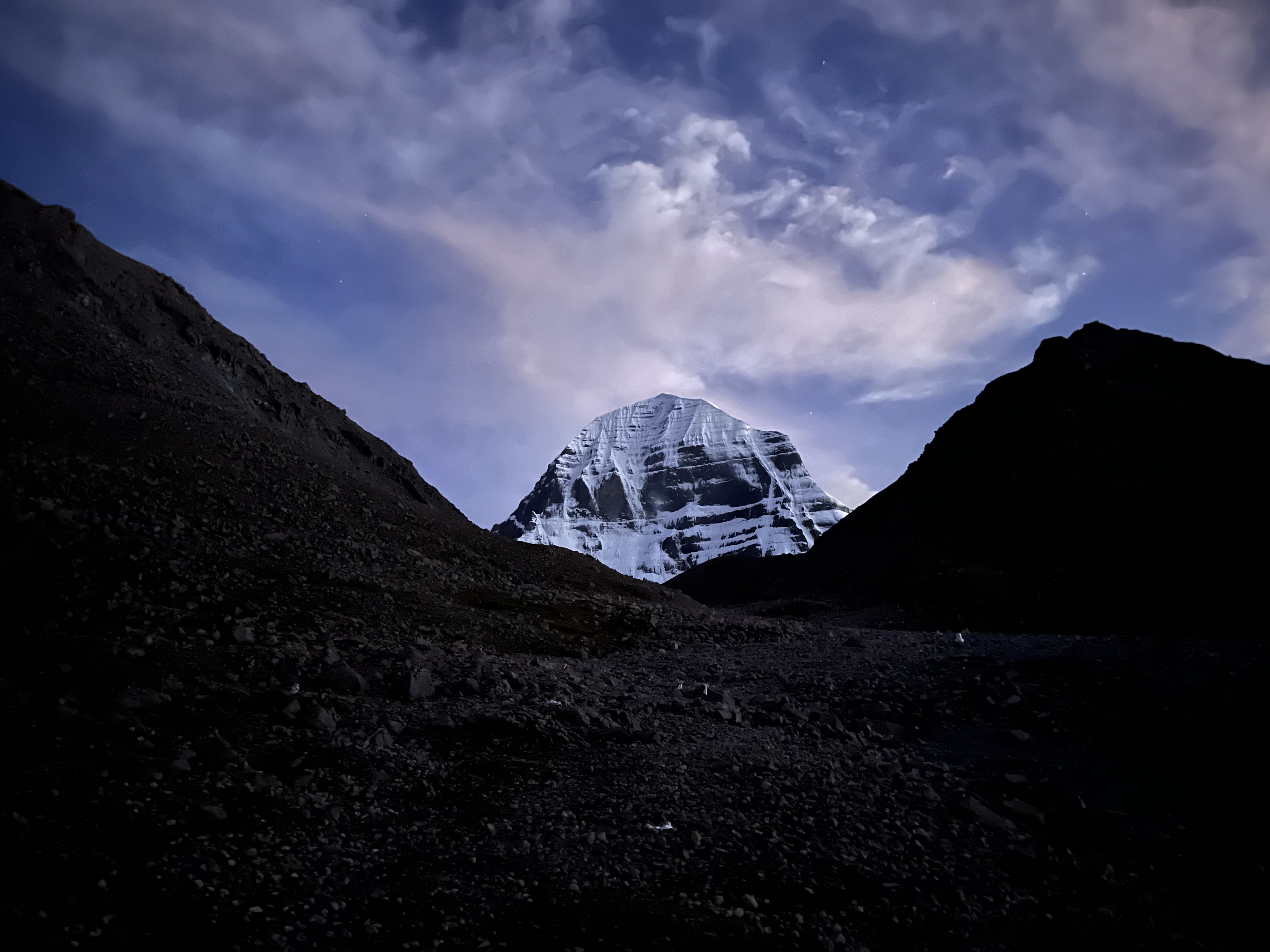 Mount Kailash at twilight, snow-lit peak framed by dark valley slopes under a starry sky
